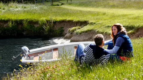 Zwei Menschen sitzen am Ufer eines Flusses neben einem Boot im Wasser.